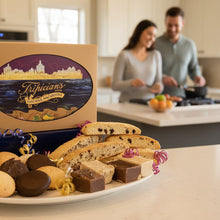 Load image into Gallery viewer, A selection of baked goods including macaroons, biscotti, and fudge arranged on a plate with a decorative bow and gift box in the background, Tripician brand